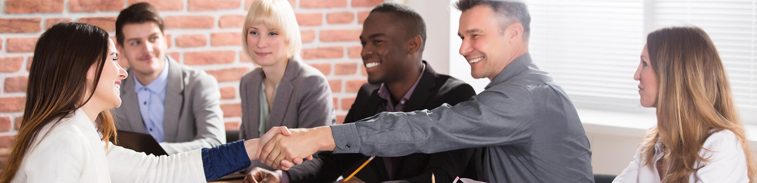 Female shakes hand of male as others the table smile.